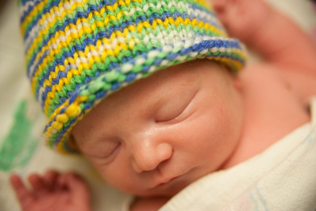A newborn baby wearing a yellow, white, blue, and green striped knit hat.